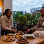Two women in cozy sweaters sharing a meal and wine on an outdoor terrace in Nairobi during June, with a live acoustic guitar player in the background.