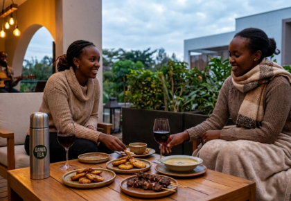 Two women in cozy sweaters sharing a meal and wine on an outdoor terrace in Nairobi during June, with a live acoustic guitar player in the background.